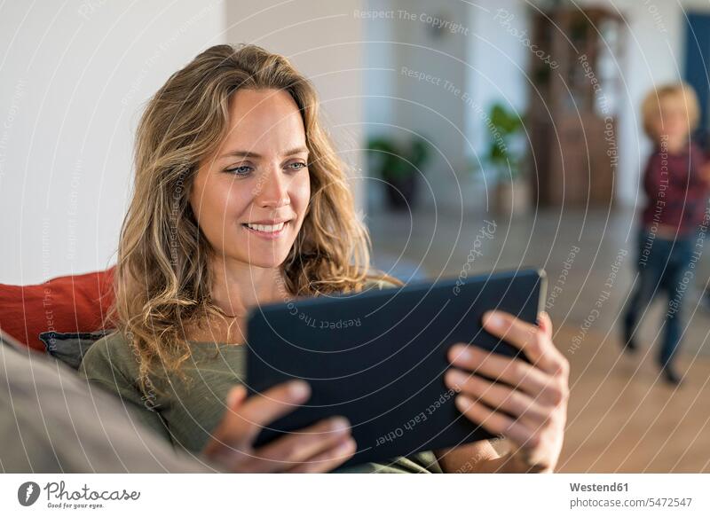 Smiling woman realxing on couch at home using tablet with son in background digitizer Tablet Computer Tablet PC Tablet Computers iPad Digital Tablet