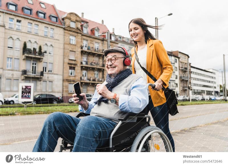 Smiling young woman pushing happy senior man with headphones and smartphone in wheelchair human human being human beings humans person persons