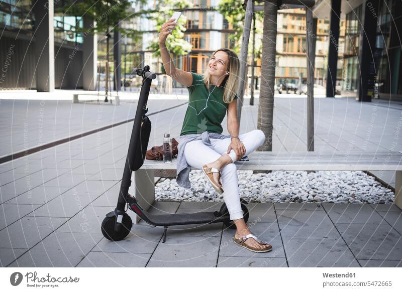 Smiling mid adult woman listening music and taking selfie while sitting on bench in city color image colour image outdoors location shots outdoor shot