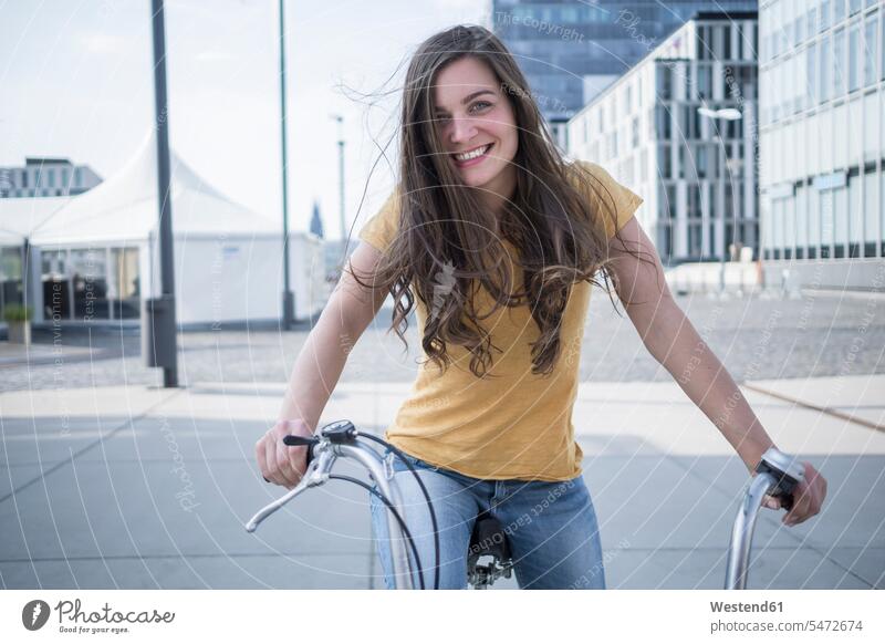 Germany, Cologne, portrait of smiling young woman with blowing hair on her bicycle caucasian european caucasian ethnicity caucasian appearance outdoors