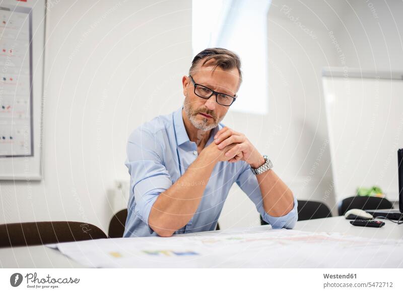 Mature businessman looking at plan on desk in office offices office room office rooms plans Businessman Business man Businessmen Business men desks eyeing