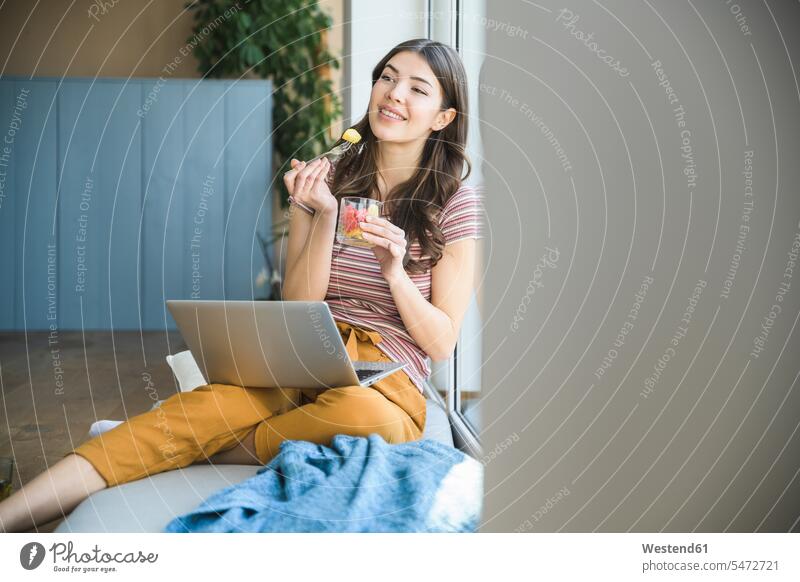 Young woman sitting at the window at home with laptop having a snack smiling smile Snack Snacks Snack Food females women Seated eating Laptop Computers laptops