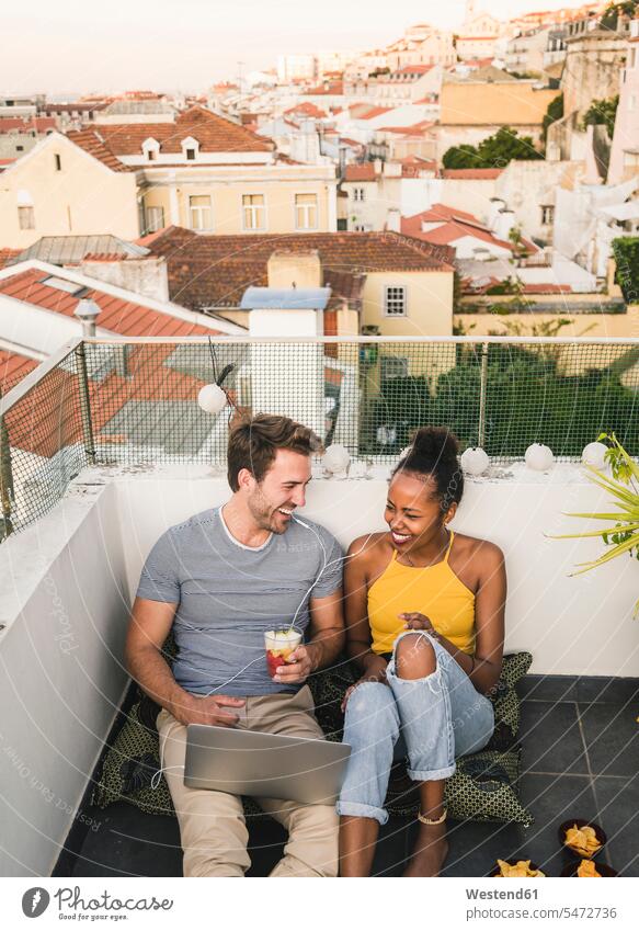 Happy young couple with laptop and earphones sitting on rooftop in the evening, Lisbon, Portugal human human being human beings humans person persons African