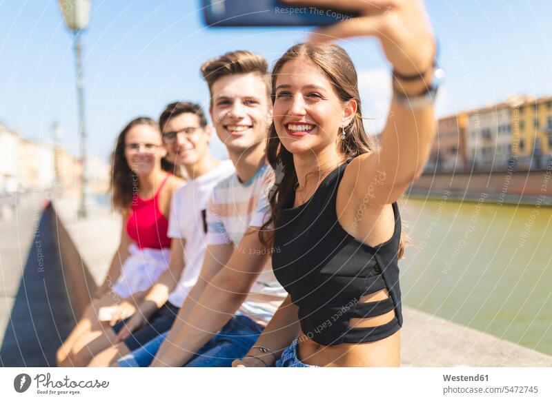 Italy, Pisa, group of four happy friends sitting on a wall along Arno river taking a selfie Seated walls Selfie Selfies together riverside riverbank happiness