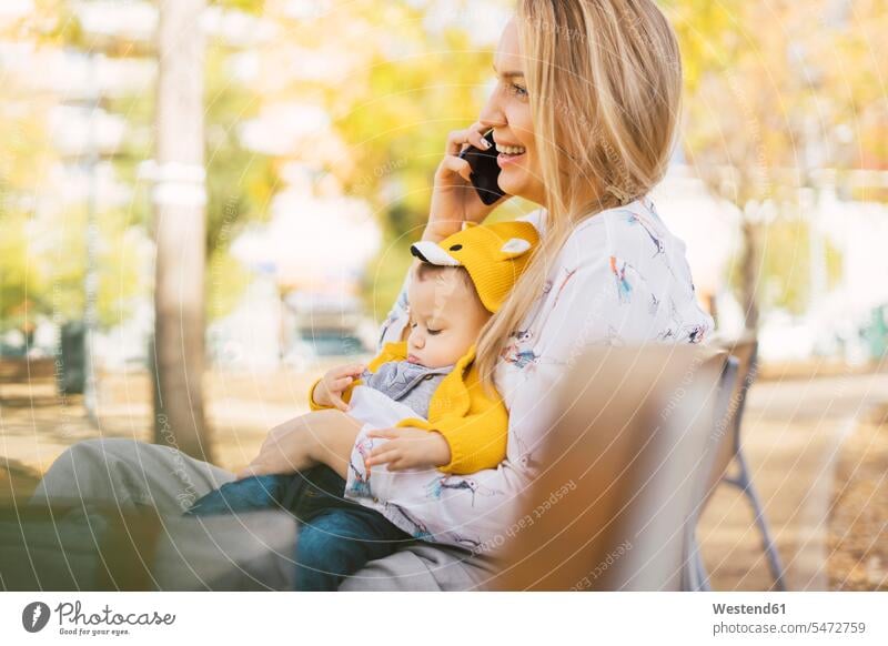 Happy mother resting with baby boy on a park bench talking on the phone coat coats jackets benches park benches telecommunication phones telephone telephones
