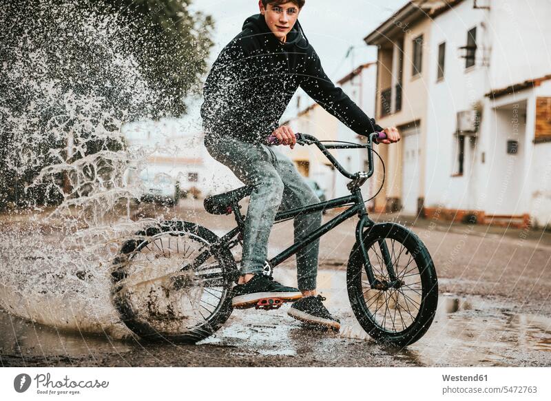 Confident teenage boy splashing water in puddle while cycling on street during rainy season color image colour image outdoors location shots outdoor shot