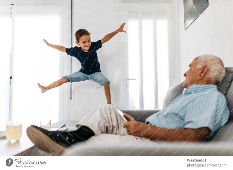 Grandfather sitting on the sofa with tablet watching his grandson jumping in the air grandsons grandfather grandpas granddads grandfathers couch settee sofas