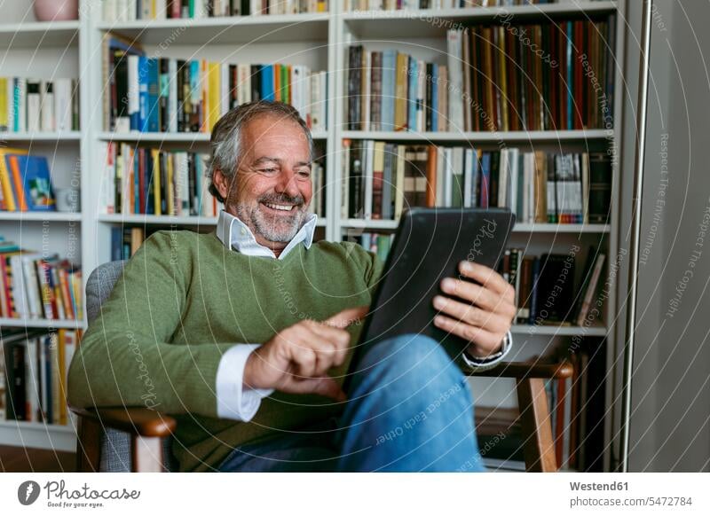 Smiling man using digital tablet while sitting against bookshelf at home color image colour image indoors indoor shot indoor shots interior interior view