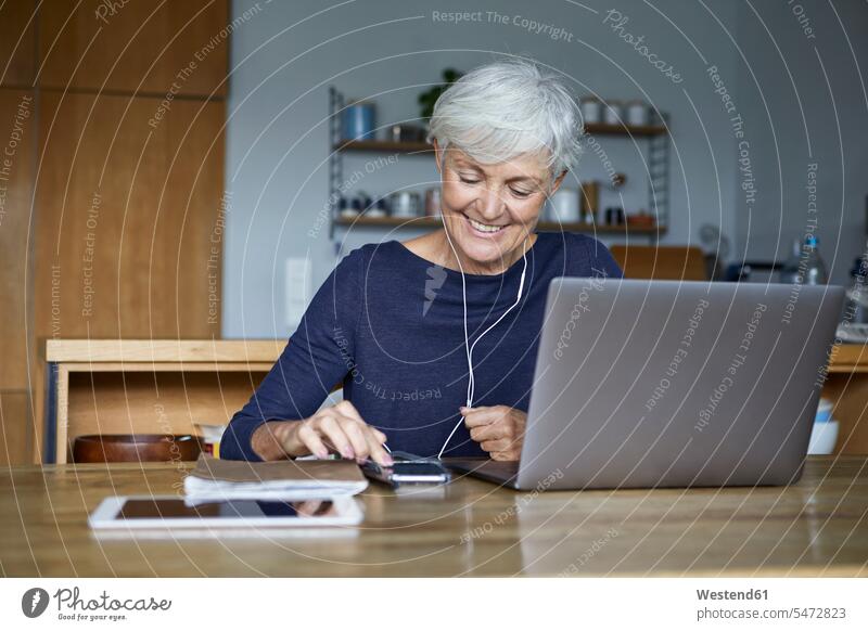 Smiling woman listening to music on smart phone and using laptop while sitting at home color image colour image indoors indoor shot indoor shots interior
