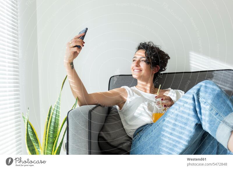 Smiling young woman sitting on couch with a soft drink taking a selfie human human being human beings humans person persons curl curled curls curly hair