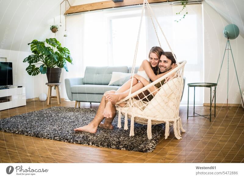 Happy affectionate couple sitting in hanging chair at home Affection Affectionate Seated happiness happy twosomes partnership couples Swinging Chair