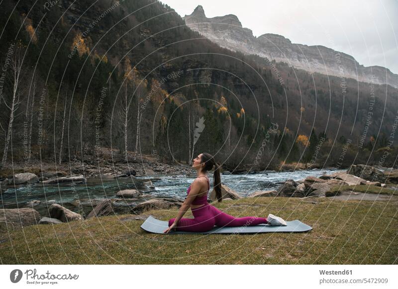 Active woman practicing yoga on mat by river and mountains at Ordesa National Park, Huesca, Spain color image colour image outdoors location shots outdoor shot