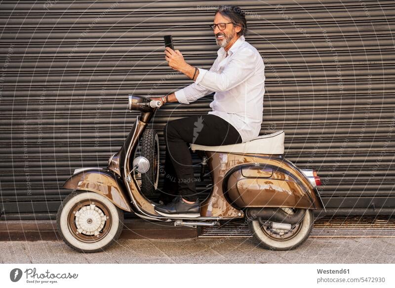 Happy mature man taking selfie while sitting on motor scooter against black shutter color image colour image outdoors location shots outdoor shot outdoor shots