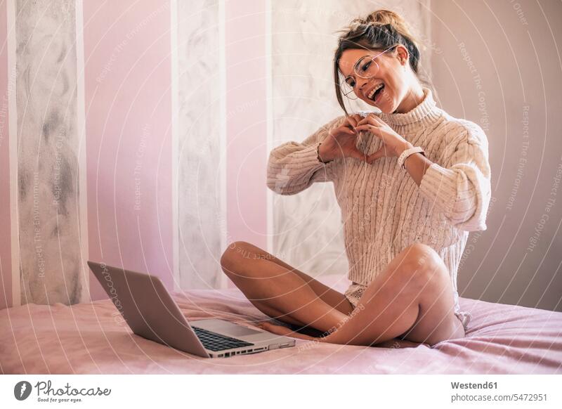 Young woman sitting on bed with laptop forming a heart with her hands during video chat beds Laptop Computers laptops notebook hearts heart shapes shaping