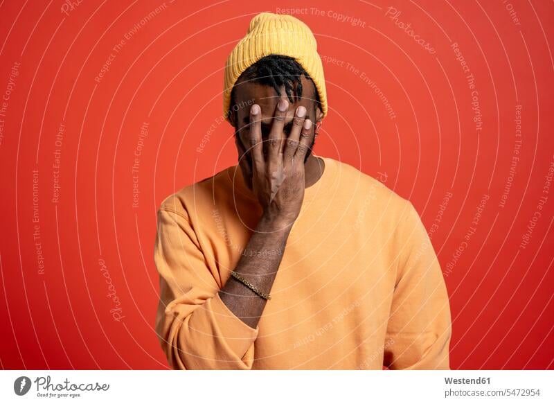 Young man covering his face in front of orange wall jumper sweater Sweaters pessimistic Emotions Feeling Feelings Sentiment Sentiments despaired desperate