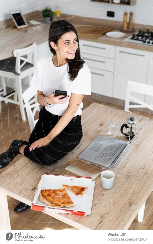 Young woman with cell phone eating pizza in kitchen at home human human being human beings humans person persons celibate celibates singles solitary people