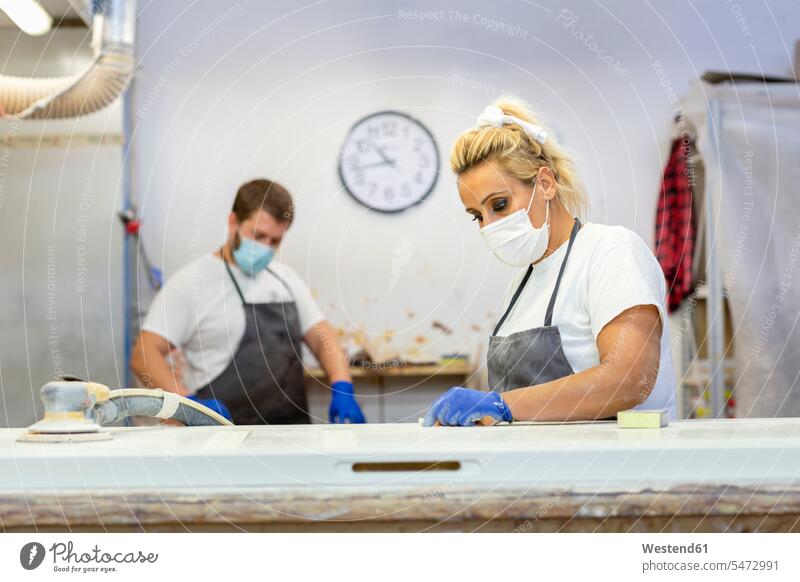 Woman with face mask working by workbench with coworker standing in background at workshop color image colour image indoors indoor shot indoor shots interior