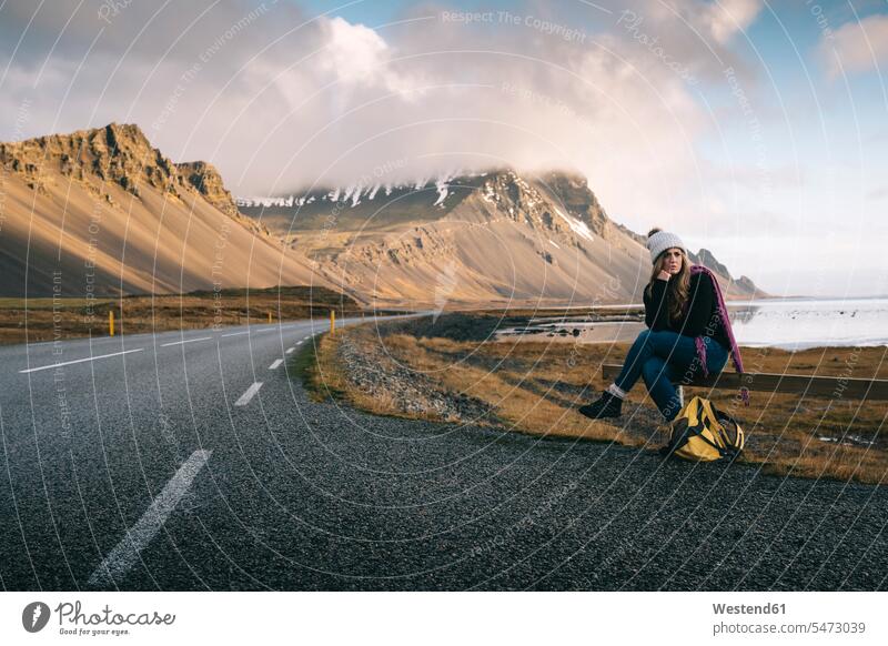 Woman with backpack sitting on railing while waiting at roadside against mountains in Iceland color image colour image outdoors location shots outdoor shot
