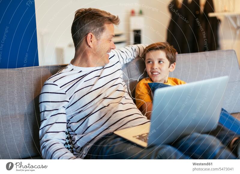 Smiling father and son with earbuds and laptop on couch at home Laptop Computers laptops notebook sons manchild manchildren settee sofa sofas couches settees