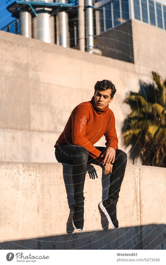 Portrait of fashionable young man wearing turtleneck pullover sitting on a wall at sunlight Seated walls Sunlit portrait portraits men males sunshine Sunny Day