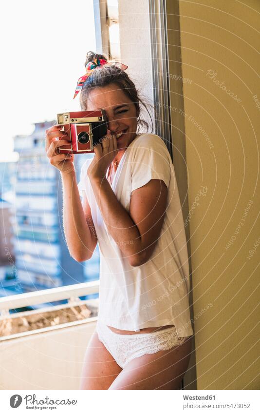 Portrait of happy young woman standing at the window taking picture with old-fashioned camera photographing t-shirt tee-shirt T- Shirt t-shirts photographer