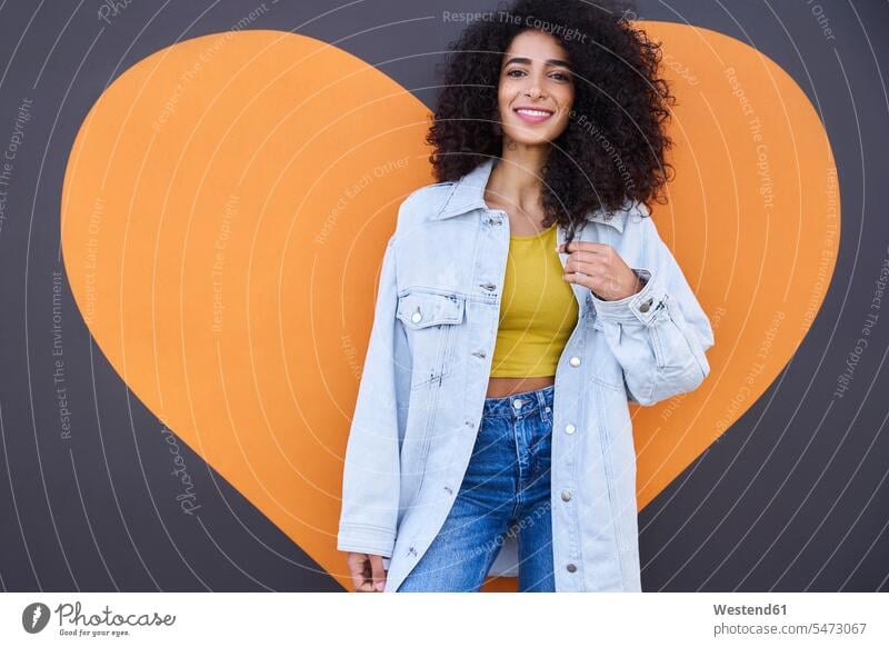 Beautiful woman with curly hair wearing denim jacket standing against heart shape on wall color image colour image Germany leisure activity leisure activities