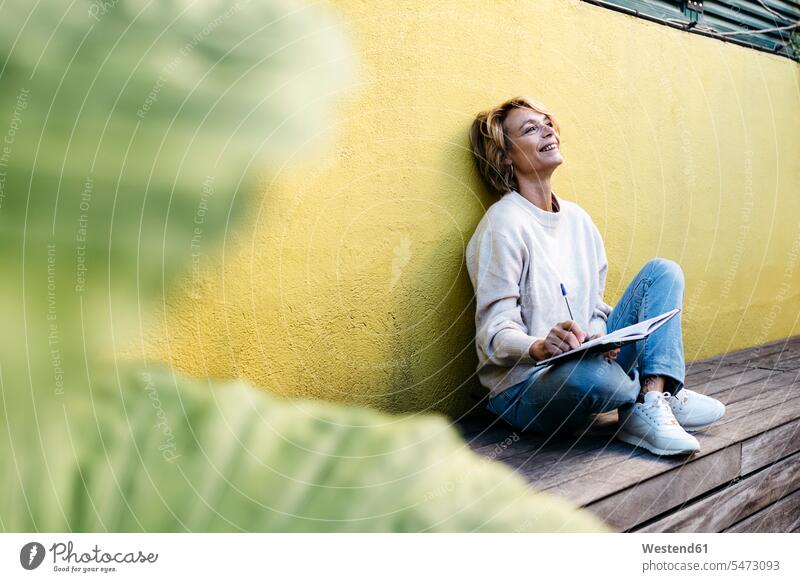 Thoughtful smiling woman sitting with diary against yellow wall on building terrace color image colour image outdoors location shots outdoor shot outdoor shots