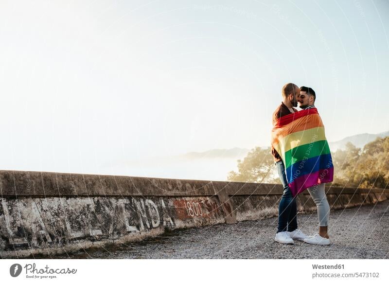 Gay couple wrapped in a gay pride flag kissing on a road in the mountains human human being human beings humans person persons caucasian appearance