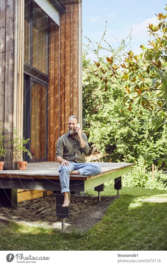 Bearded mature man talking over mobile phone while sitting outside tiny house color image colour image Germany leisure activity leisure activities free time