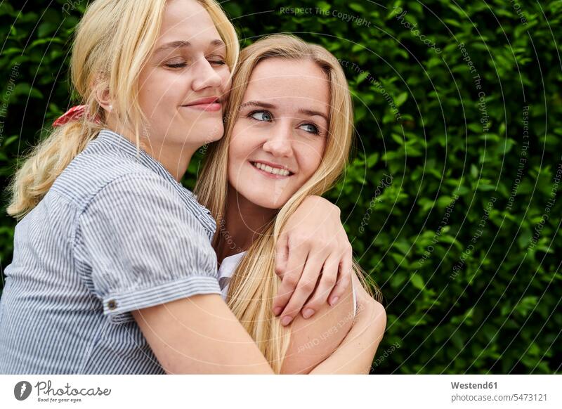 Two happy young women hugging a hedge embracing embrace Embracement hedges Hedgerow woman females female friends happiness Adults grown-ups grownups adult
