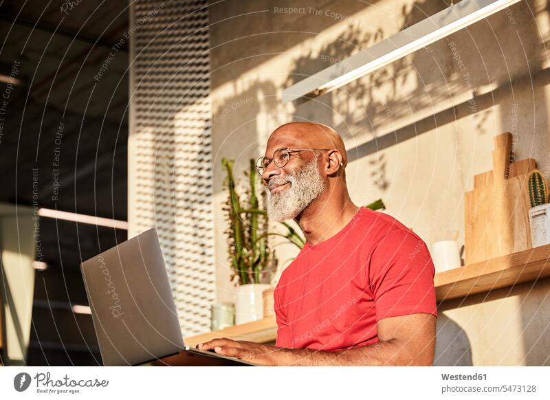 Smiling mature man looking away while using laptop at home on sunny day color image colour image indoors indoor shot indoor shots interior interior view