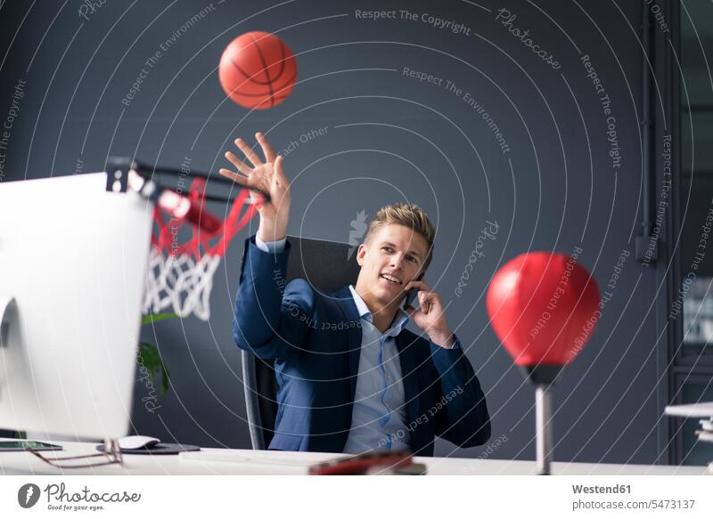 Young businessman sitting at desk in office playing basketball and talking on cell phone Businessman Business man Businessmen Business men offices office room