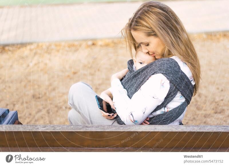 Happy mother resting with baby boy on a park bench benches park benches hold cuddle snuggle snuggling smile Seated sit embrace Embracement hug hugging relax