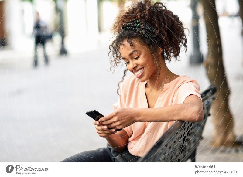 Smiling young woman sitting on a bench using cell phone females women Seated happiness happy mobile phone mobiles mobile phones Cellphone cell phones benches