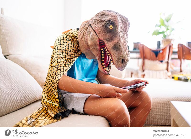 Boy wearing dinosaur mask and cape using smart phone while sitting on sofa at home color image colour image Spain indoors indoor shot indoor shots interior