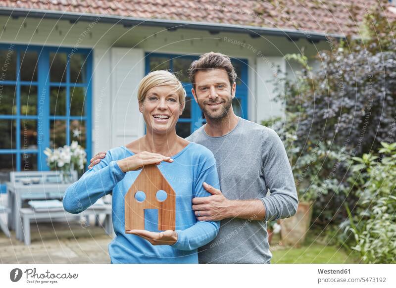 Portrait of smiling couple standing in front of their home holding house model models portrait portraits houses twosomes partnership couples smile building