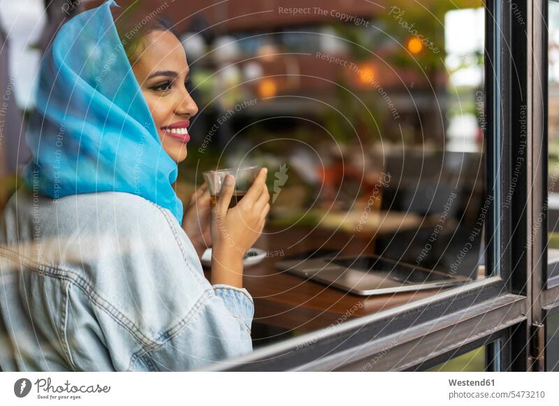 Young woman wearing turquoise hijab and drinking tea in a cafe, looking through window windows head cloth head cloths head scarf head scarves headscarves
