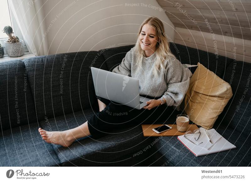 Smiling freelancer using laptop while sitting on sofa with drink and document at home color image colour image indoors indoor shot indoor shots interior