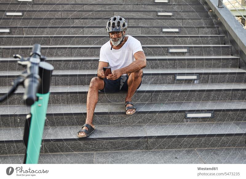 Smiling mature man with E-Scooter sitting on stairs using cell phone human human being human beings humans person persons Mixed Race mixed race ethnicity