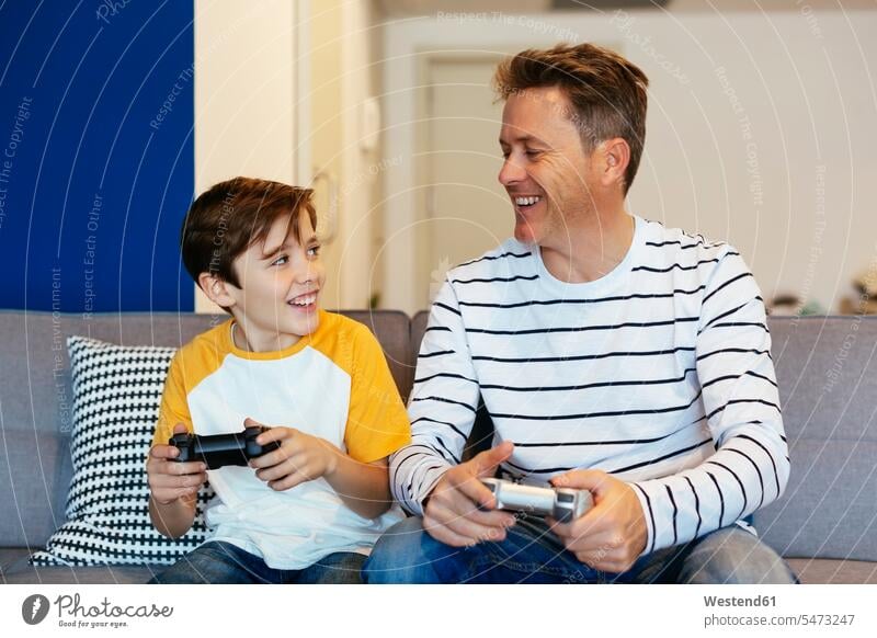 Happy father and son playing video game on couch at home settee sofa sofas couches settees pa fathers daddy dads papa video games sons manchild manchildren