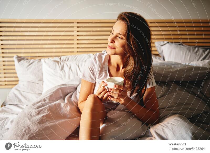 Portrait of smiling young woman sitting on bed with cup of coffee looking at distance beds smile view seeing viewing Seated Coffee females women Drink beverages