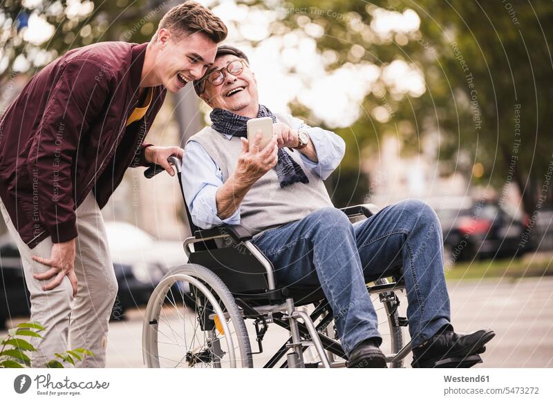 Laughing senior man in wheelchair and his adult grandson looking together at smartphone having fun generation phones telephone telephones cell phone cell phones