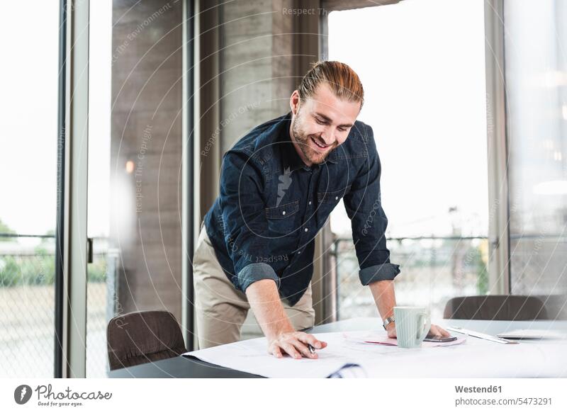 Smiling young businessman working on plan at desk in office At Work plans offices office room office rooms smiling smile desks Businessman Business man
