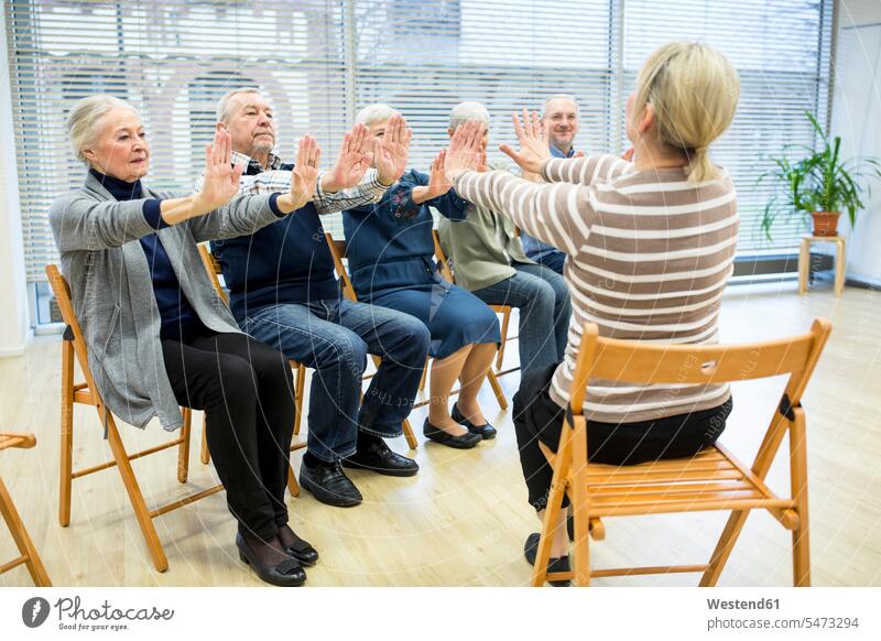 Group of seniors practicing chair gymnastics with instructor in retirement home chairs Seated sit exercise exercising practice practise practising Retired