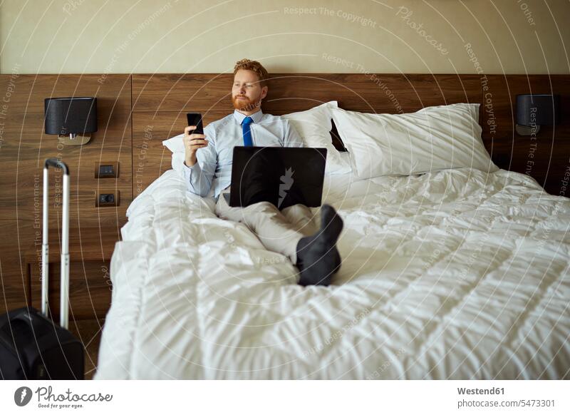 Businessman lying on bed in hotel room using laptop and cell phone Occupation Work job jobs profession professional occupation business life business world
