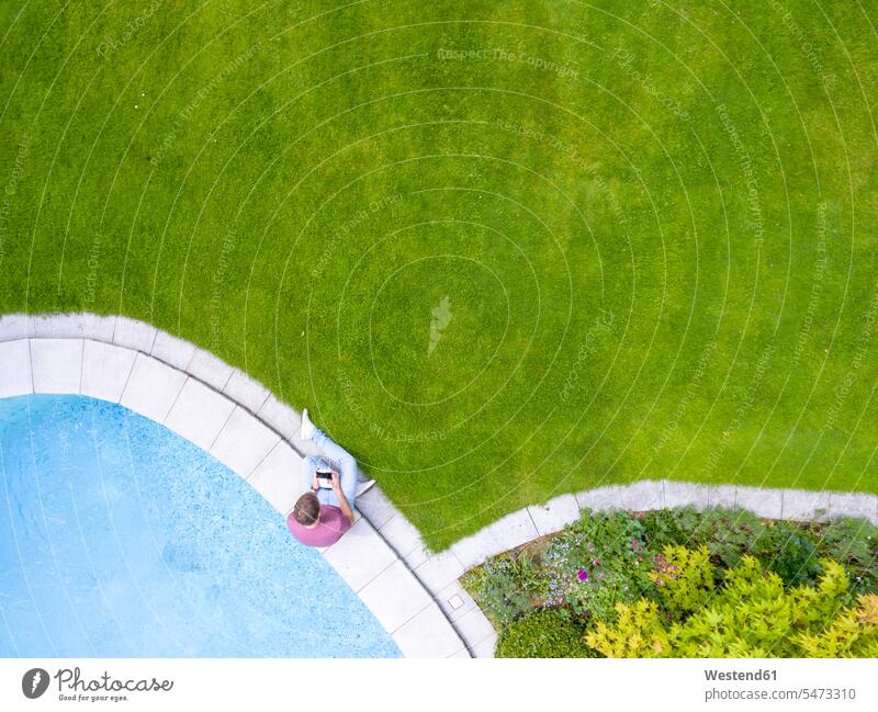 Man sitting at poolside with remote control navigating drone navigate man men males pool edge Pool Side drones swimming pool swimming pools Seated