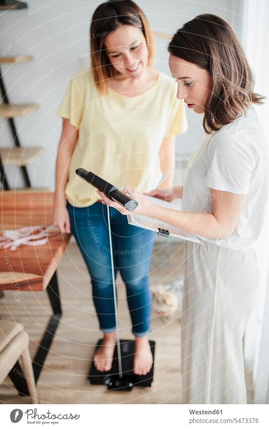 Young woman measuring friend's weight on weight scale at home color image colour image indoors indoor shot indoor shots interior interior view Interiors day