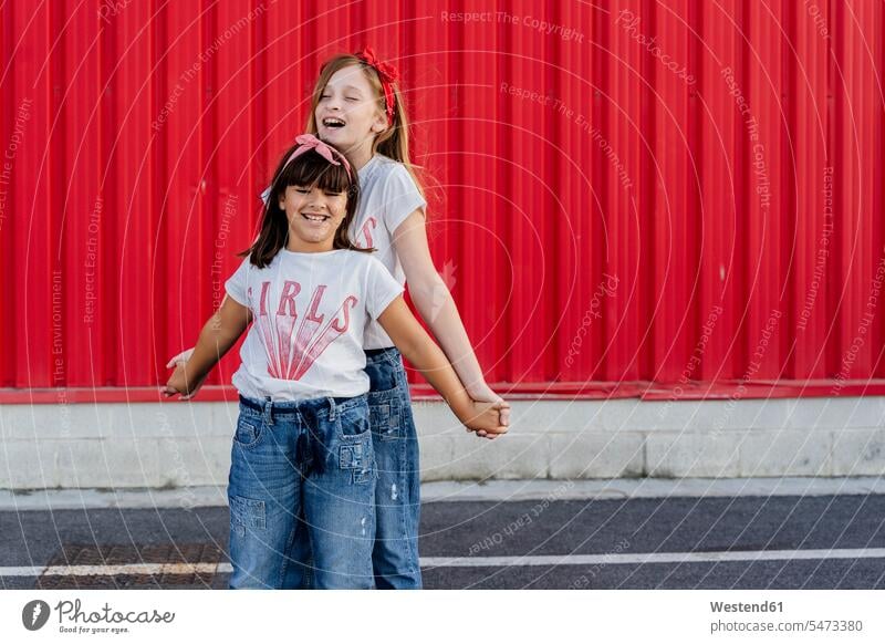Sisters standing in front of a red wall pants Trouser Denim Jeans T- Shirt t-shirts tee-shirt smile delight enjoyment Pleasant pleasure indulgence indulging