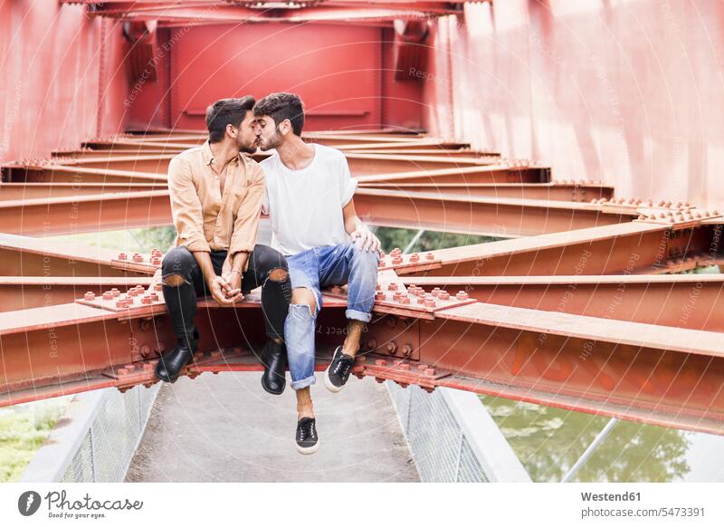 Kissing young gay couple sitting on steel girder of a footbridge Seated kissing kisses twosomes partnership couples gay men gay man homosexual men