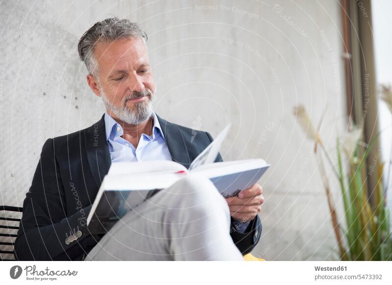 Smiling businessman sitting at concrete wall reading book Seated books concrete walls Businessman Business man Businessmen Business men smiling smile